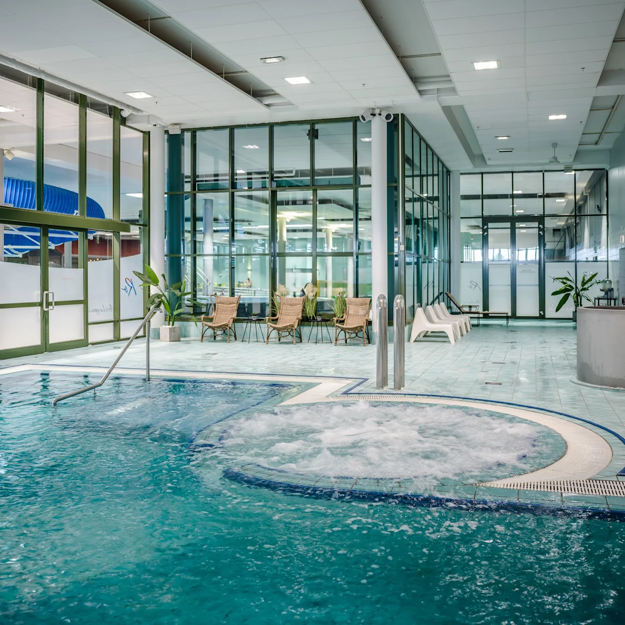 Swimming pool with bubbling water, surrounded by chairs and plants, inside a glass-walled recreational facility. Blue water slide visible outside through glass doors. Signs in a Scandinavian language.