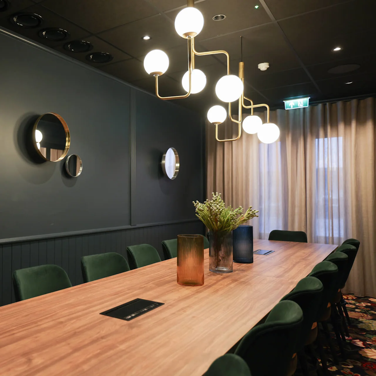 A wooden table set with vases and chairs occupies a room with dark walls. Decorative round mirrors and modern lights adorn the walls, with curtains covering the windows.