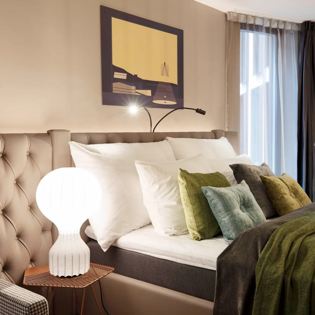Bed adorned with white and green pillows sits beside a small side table holding a white lamp. A modern painting hangs above, while a window with sheer curtains illuminates the room.