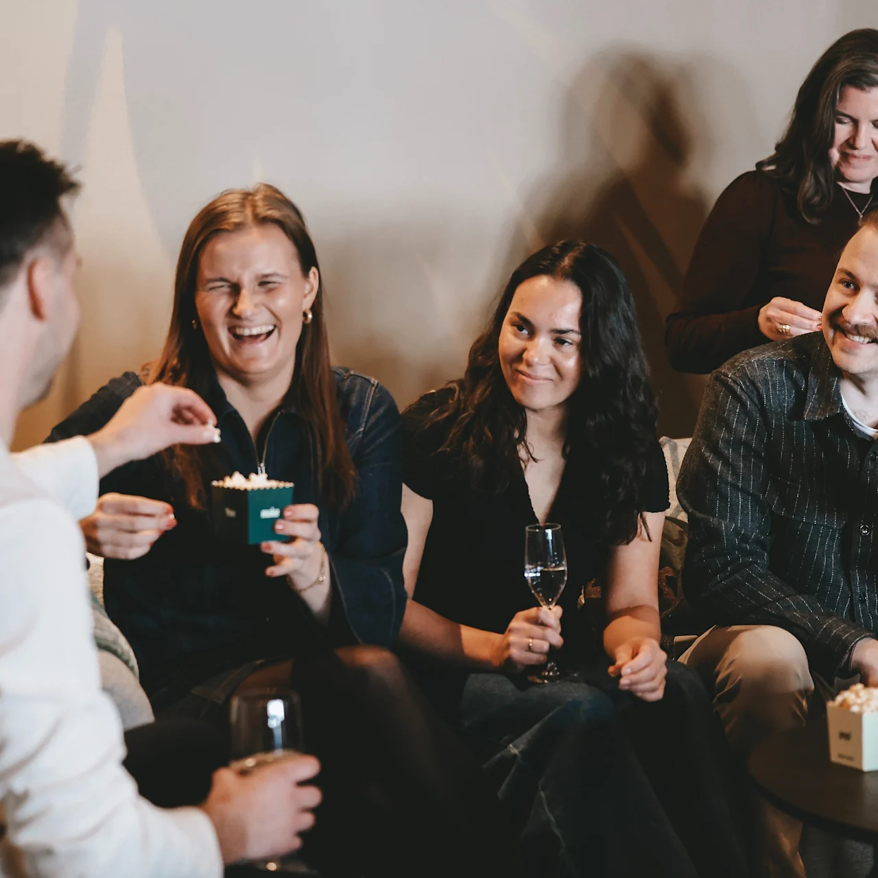 A group of five people sits and stands while smiling and chatting in a cozy room. Some hold drinks and small boxes, suggesting a relaxed social gathering.