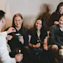 A group of five people sits and stands while smiling and chatting in a cozy room. Some hold drinks and small boxes, suggesting a relaxed social gathering.