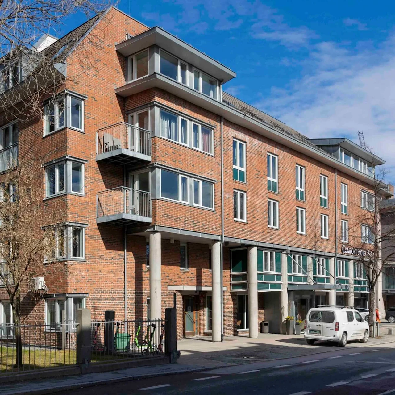 Monikerroksinen tiilitalo parvekkeineen seisoo tien vieressä sinisen taivaan alla. Valkoinen pakettiauto on pysäköity läheisyydessä. Kyltti lukee "Comfort Hotel Børsparken."