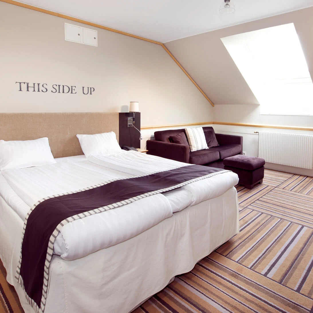 Ett välordnat hotellrum med dubbelsäng, soffa och skrivbord under snedtak. Text på väggen lyder "THIS SIDE UP".