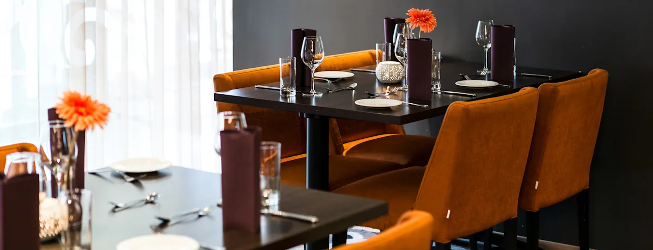 Ruokapöytä katettu laseilla, lautasilla ja lautasliinoilla; koristeltu oransseilla kukilla hämärästi valaistussa ravintolassa; seinällä muotokuva.