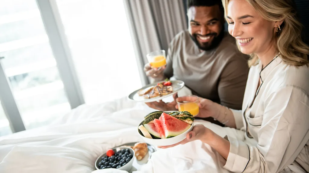 A smiling couple enjoying breakfast in bed in a hotel room.