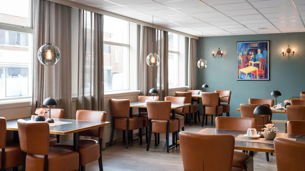 Bord och stolar står i en tom, väl upplyst restaurang. Läderstolar, hängande lampor och en färgglad målning på väggen definierar den eleganta miljön.