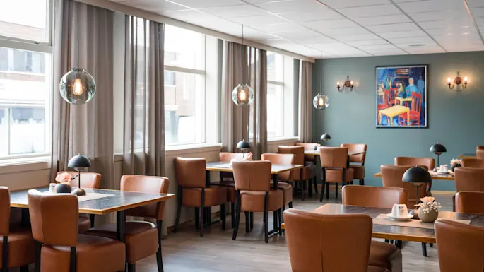 Tables and chairs sit in an empty, well-lit restaurant. Leather chairs, pendant lights, and a colorful painting on the wall define the elegant setting.