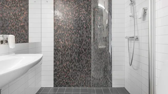 Ett badrum har en dusch med ett modernt duschmunstycke på en vit kaklad vägg. Bakväggen är dekorerad med små svarta mosaikplattor. En vit vask är bredvid.