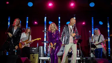 Et livlig band opptrer på scenen under sterke lys, med musikere som spiller gitar og sangere som smiler og engasjerer publikum.