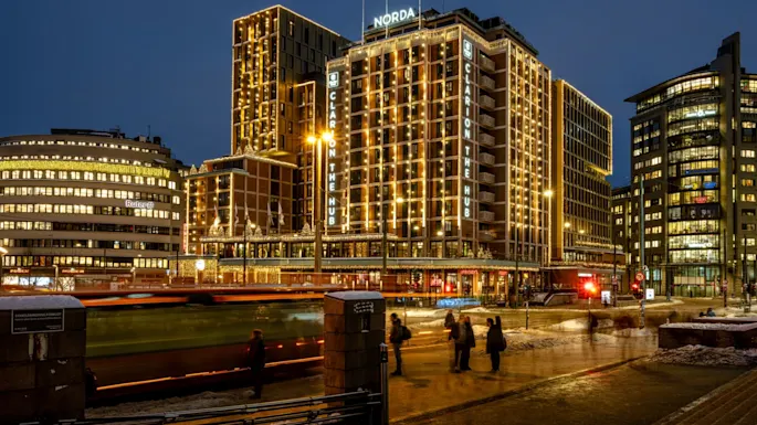 A tall, illuminated hotel building, decorated with lights, stands beside a busy street where a bus passes and people walk. Text includes: "Ruter", "Norda", "Clarion The Hub", "TGI Friday's".