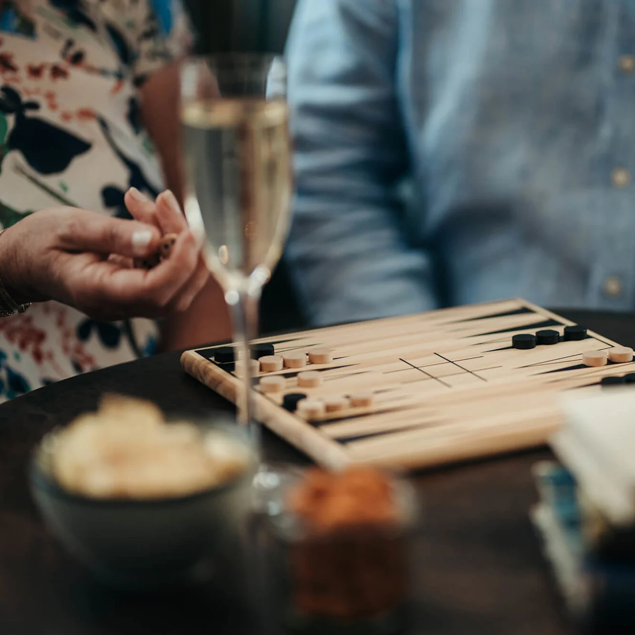 Two people are playing backgammon at a cozy table with drinks and snacks.