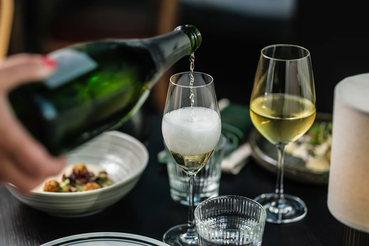 En grön flaska häller mousserande vin i ett glas, skapar skum. Bredvid finns ett fullt vinglas. Tallrikar och en servett syns på ett mörkt bord.