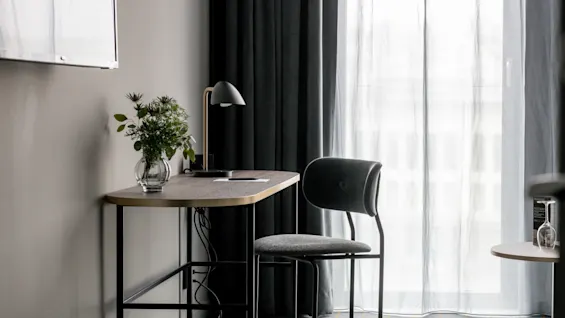 Et lille skrivebord med en vase med blomster og en sort lampe står ved siden af en mørk stol, tæt på let gennemsigtige gardiner, der lader lys komme ind i rummet.