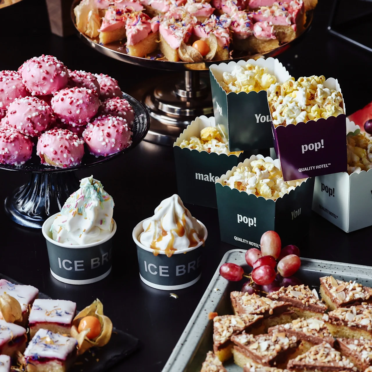 Pink pastries rest on a glass stand; next to them are popcorn in labeled containers, ice cream cups, and assorted fruits on a dark table. Containers read: "pop!", "ICE BRI", "make!", "You! QUALITY STREET".