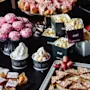 Pink pastries rest on a glass stand; next to them are popcorn in labeled containers, ice cream cups, and assorted fruits on a dark table. Containers read: "pop!", "ICE BRI", "make!", "You! QUALITY STREET".