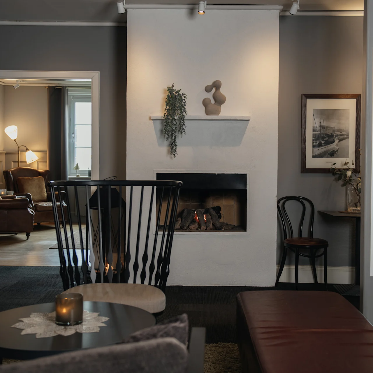 A cozy lounge features a lit fireplace in the center with elegant chairs around it. Adjacent rooms extend the seating area in a warmly lit, contemporary design.