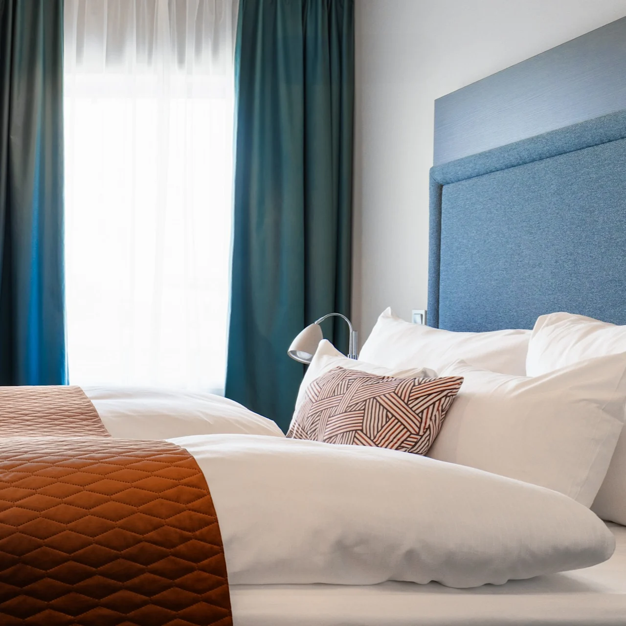 A neatly made bed with white pillows and a brown quilted runner; positioned near a large window with green curtains, creating a cozy ambiance.