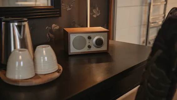 Puinen radio Bluetoothilla, vedenkeitin ja kaksi kuppia tummalla työpöydällä, vasten kuvioitua tapettia ja peiliä.