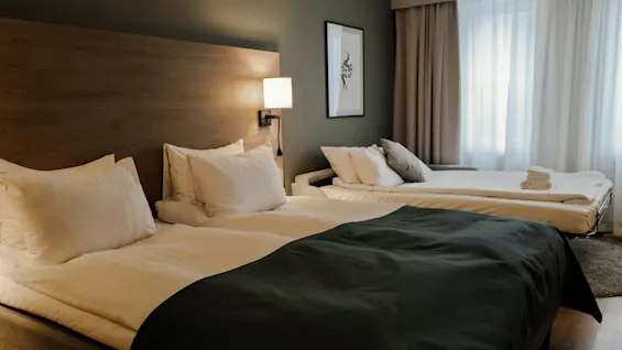 A double bed with white pillows and a dark blanket stands in a hotel room near a second bed. A lamp, picture frame, and curtains decorate the space.