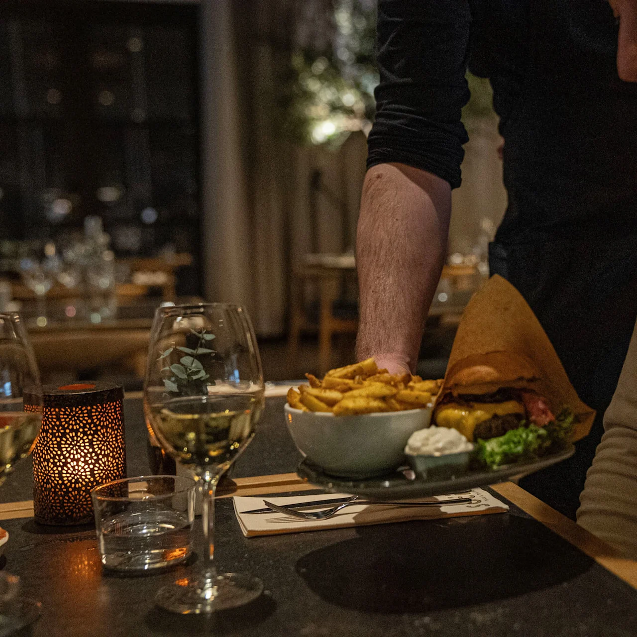 A person serves a plate of burger and fries to a seated diner in a dimly lit restaurant, with wine and a candle on the table.