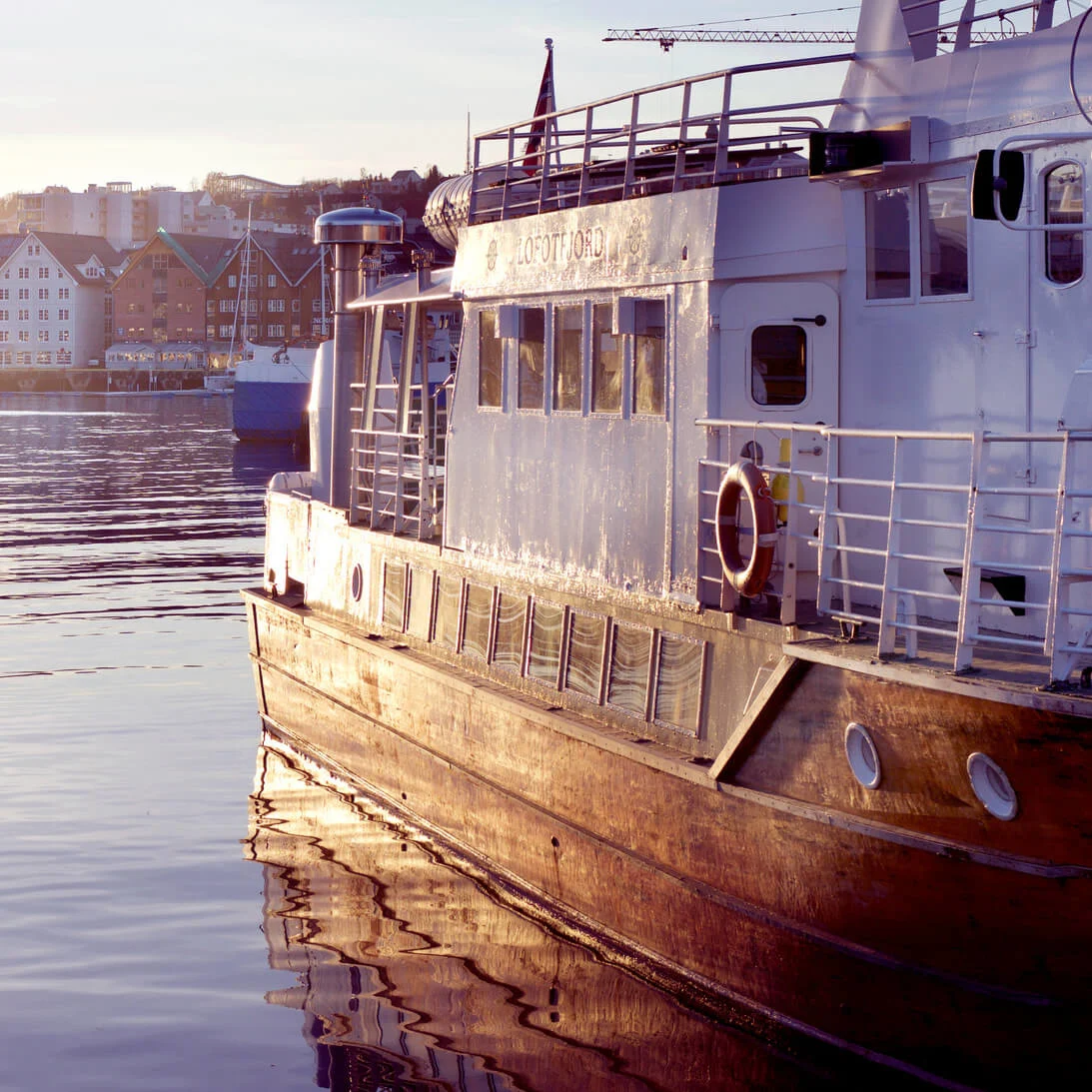 Fortøjet båd i solnedgang, gyldent lys reflekteres på roligt vand, byens silhuet i baggrunden.