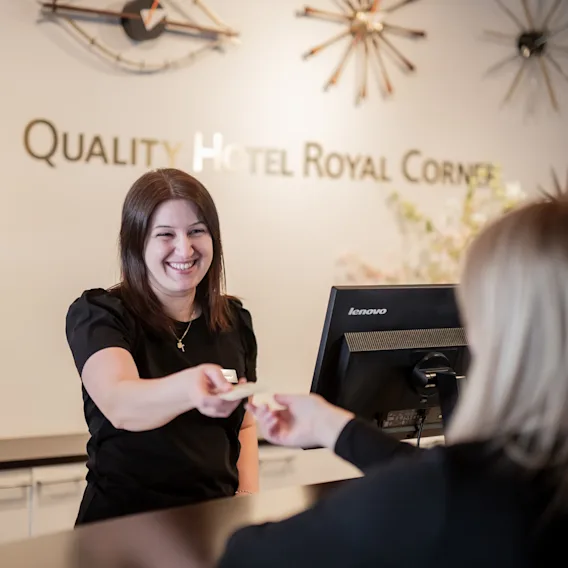 En leende receptionist ger ett kort till en gäst vid en disk. Väggen dekorerad med "QUALITY HOTEL ROYAL CORNER" text syns bakom.