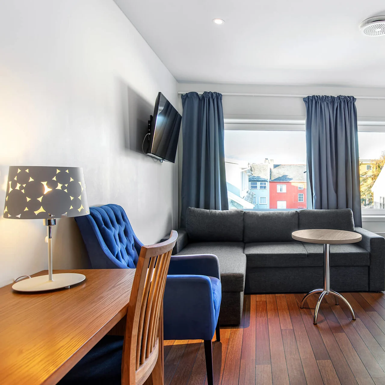 En grå sofa står mot en vegg, ved siden av et rundt bord i et moderne rom. En TV er montert over, og blå gardiner rammer inn store vinduer som viser bygninger utenfor.