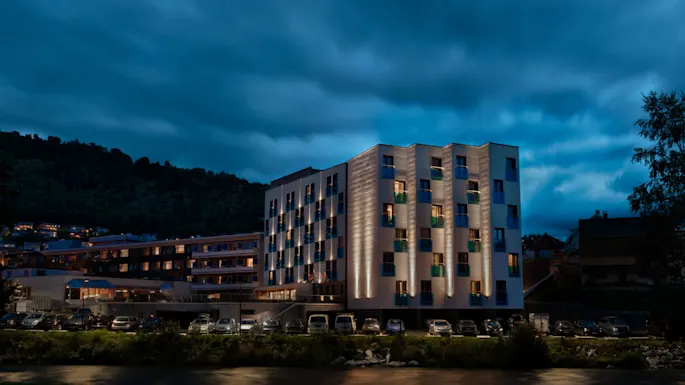 A modern five-story building with illuminated windows stands beside parked cars at night, surrounded by hills and under a cloudy sky.