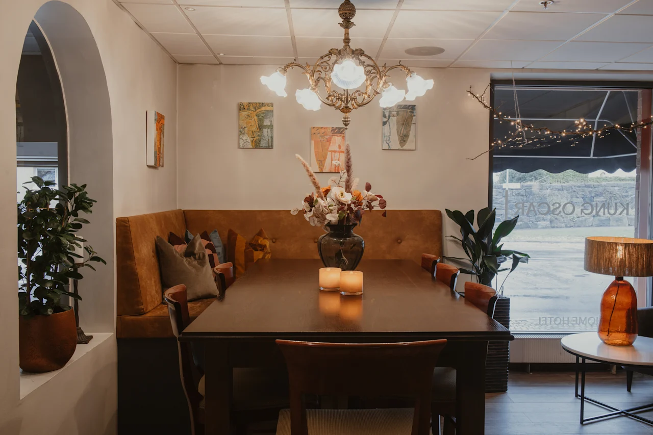 A cozy dining area at Home Hotel Kung Oscar with a long table, bench seating, floral decor, and a chandelier.