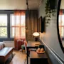 Et hyggeligt hotelværelse har en seng og et skrivebord; store vinduer med tynde gardiner lader naturligt lys strømme ind. Et spejl reflekterer en del af rummet og udsigten udenfor.