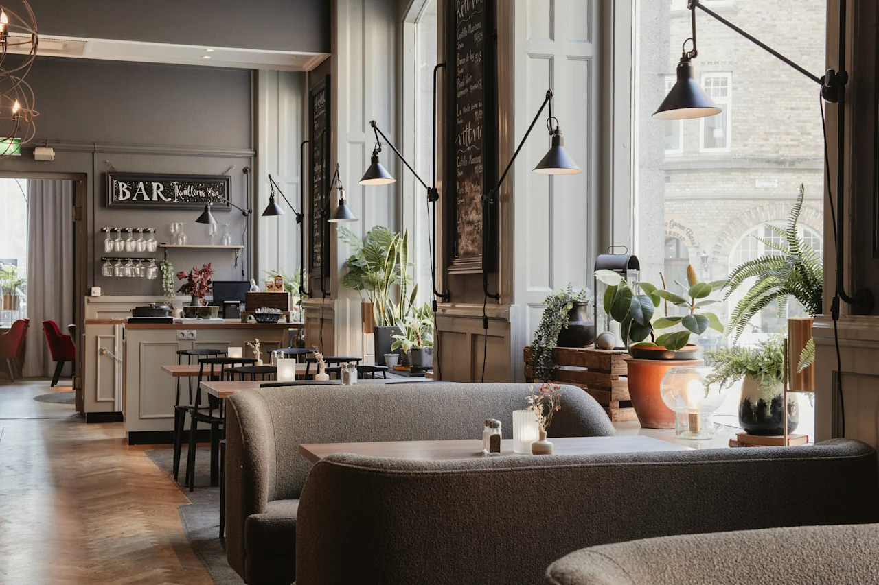 Et varmt og innbydende restaurantinteriør med komfortable båser, bord og en bar. Sollys fyller rommet og fremhever ulike planter. BAR Kvallens Vin.