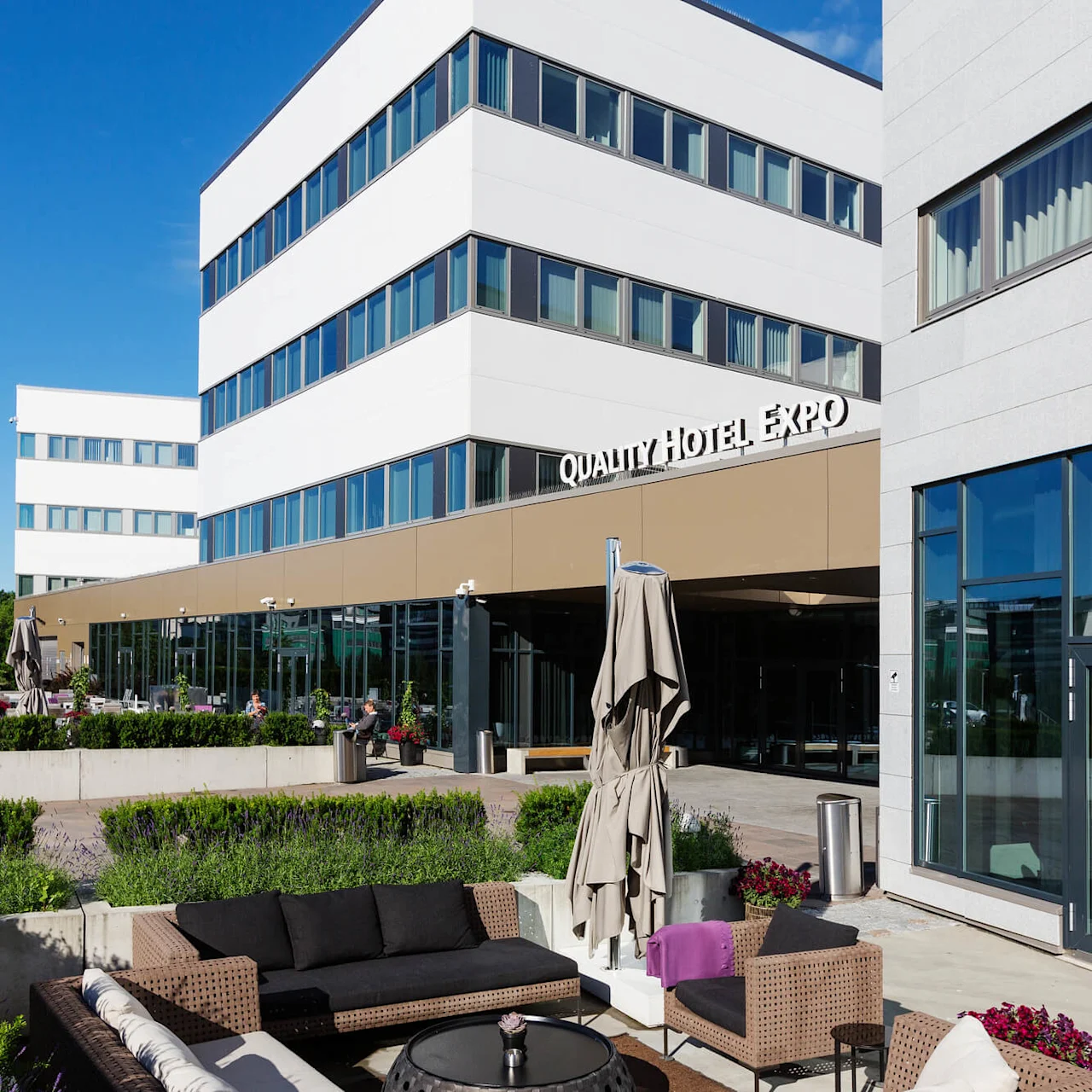 Quality Hotel Expo building with modern exterior, featuring glass and concrete. Outdoor seating area with wicker furniture and umbrellas. Surrounding greenery and clear blue sky. 