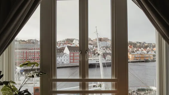 En malerisk udsigt fra et vindue over en havn med både og en charmerende by langs vandet.