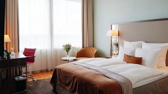 A bed with white linens and brown throw is in a hotel room, accompanied by a desk, chair, lamp, and window with sheer curtains.