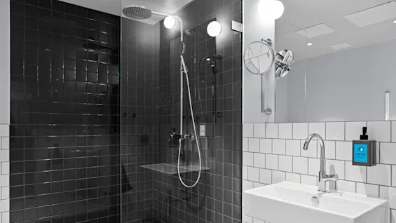 Dusjkabin med mørke fliser, med en regndusjhode og håndholdt sprøyte, i et moderne bad med hvit flislagt servantsone. Veggmontert såpe sier: "HAND WASH."