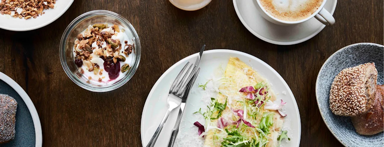 En tallerken med en omelett toppet med grønnsaker og radicchio ligger på et trebord ved siden av en kopp kaffe, et glass juice, yoghurt med granola og et rundstykke.