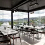 En moderne restaurant med gulv-til-tak-vinduer som tilbyr en naturskjønn utsikt over en by, elv og fjell. Bordene er dekket for bespisning.