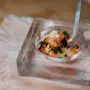 En rund glasskål i en klar blok indeholder en farverig gourmetret med urter og teksturerede pynt, præsenteret på en lys træoverflade.