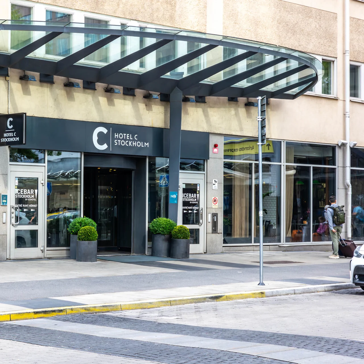 Hotel entrance displaying "HOTEL C STOCKHOLM" and "ICEBAR STOCKHOLM BY ICEHOTEL". Glass doors with potted plants, situated on a city sidewalk, with people and vehicles nearby.
