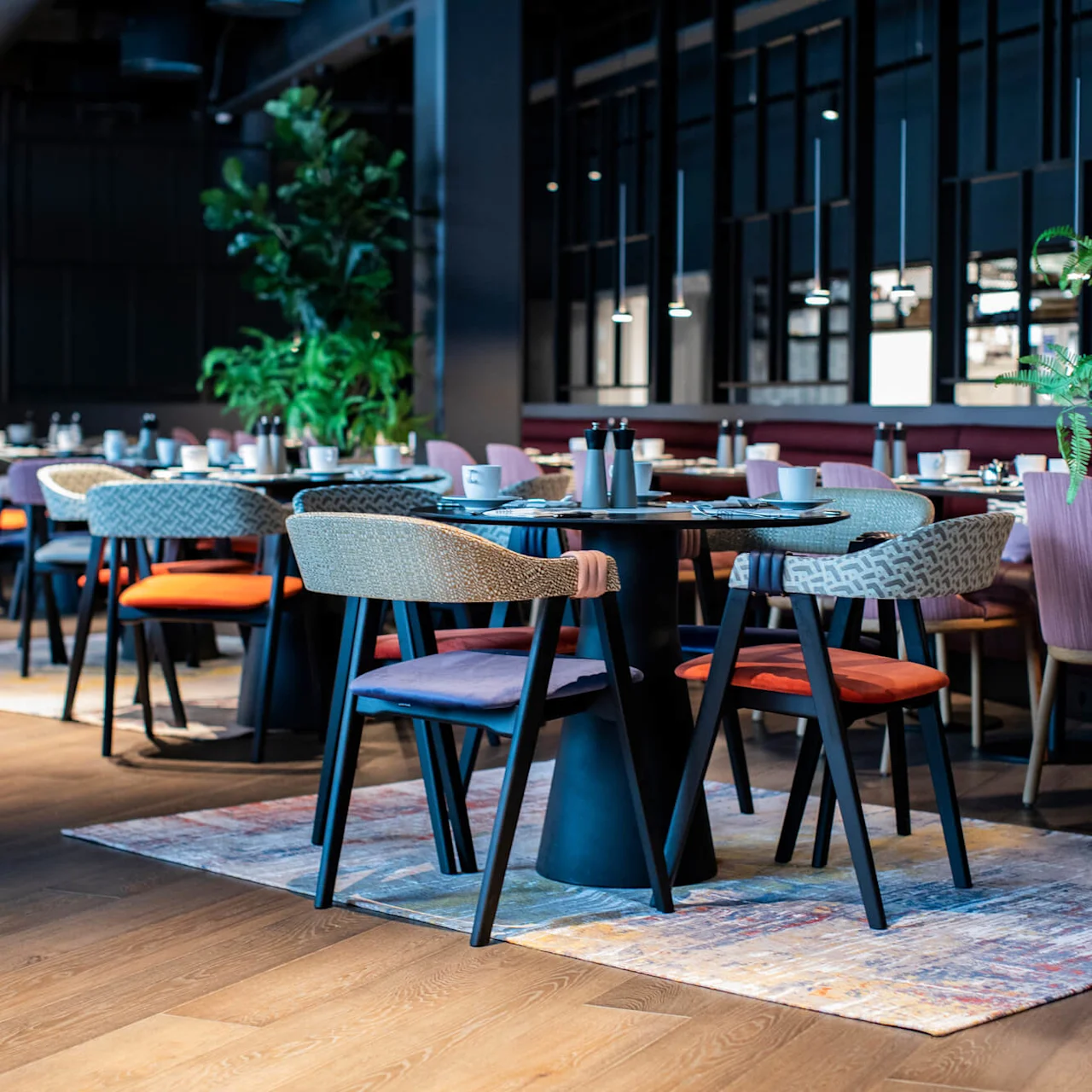 Tables and chairs arranged in a modern restaurant; tables set with cups and cutlery; surrounded by vibrant green plants and stylish lighting.