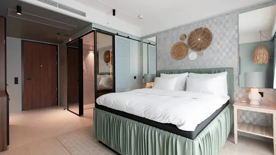 A bed with white linens stands in a modern hotel room; decorative wall art and a nightstand with a lamp add elegance.
