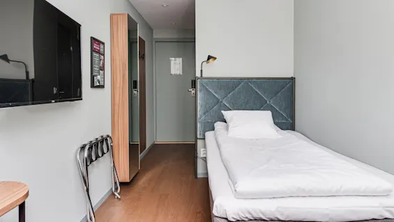A single bed with white bedding in a tidy, modern room, with a wall-mounted TV, small table, and hangers.