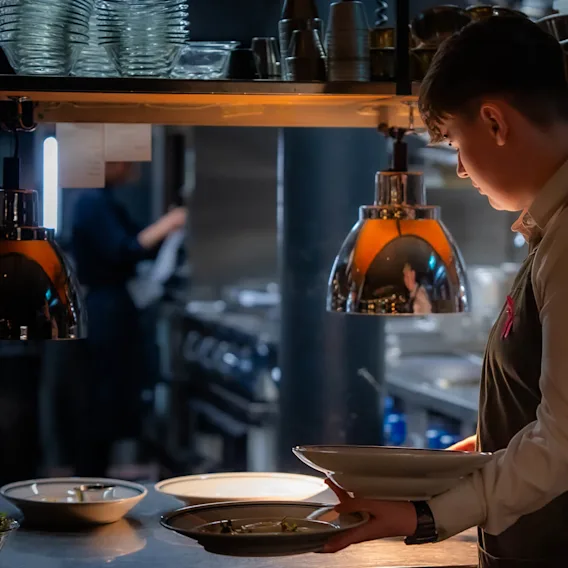 A server, wearing an apron, carries plates under warm lighting in a busy restaurant kitchen filled with shelves, metal lamps, and kitchenware.