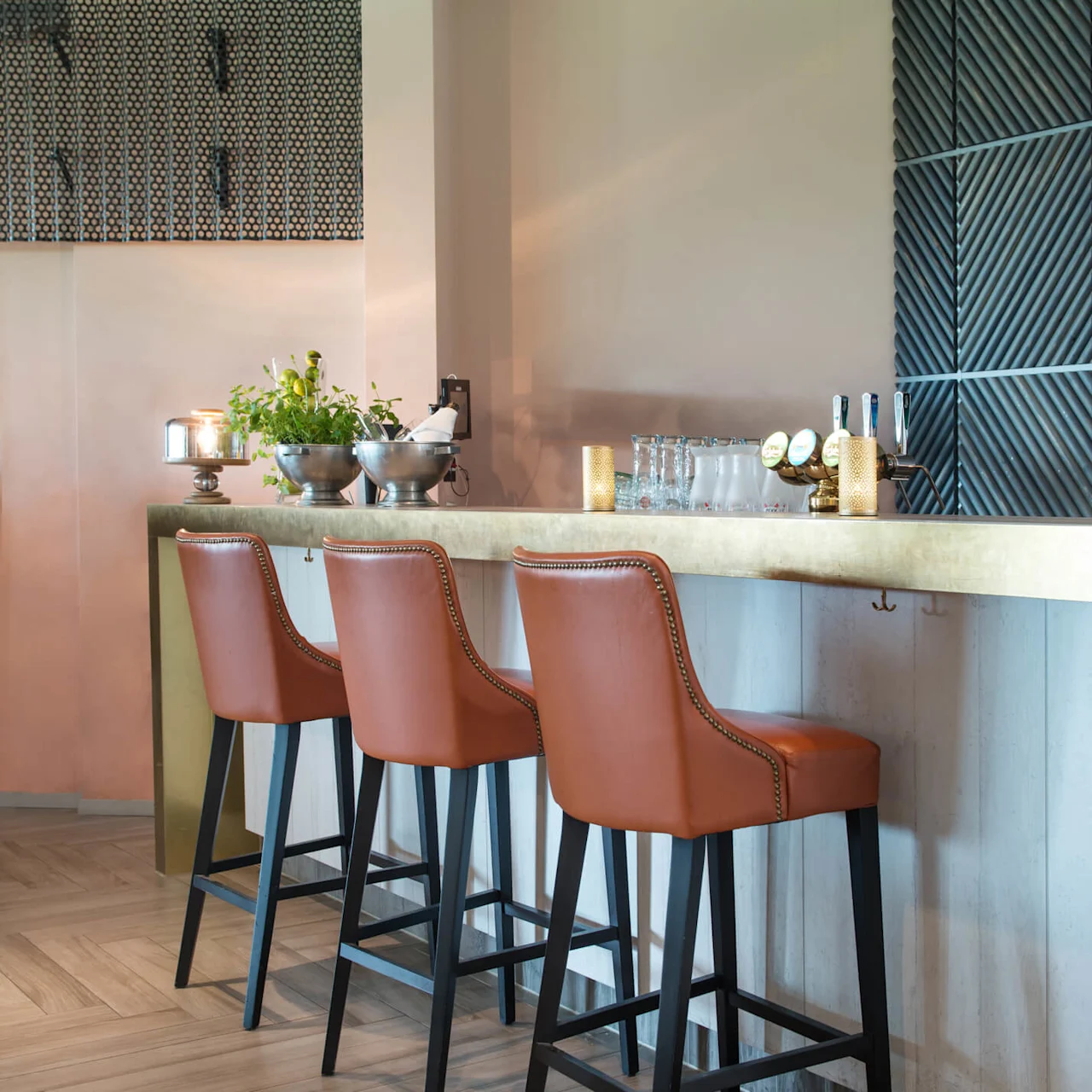 Three brown leather barstools line a bar counter with bowls of herbs and bottles, set in a modern, well-lit space with wood-patterned flooring and textured walls.