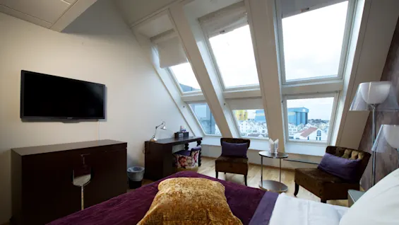 Et soverom med en stor seng, møbler, og skrå vinduer som gir utsikt over byen.
