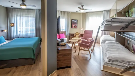 A room with a double bed and teal bedding connects to a living area through an open doorway. The living area has a pink chair, bunk beds, and wooden flooring.