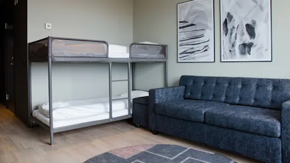 Våningssängar med vita sängkläder är placerade mot en ljusgrå vägg. En mörkblå soffa står bredvid, med två abstrakta målningar ovanför. Ljust trägolv kompletterar rummet.