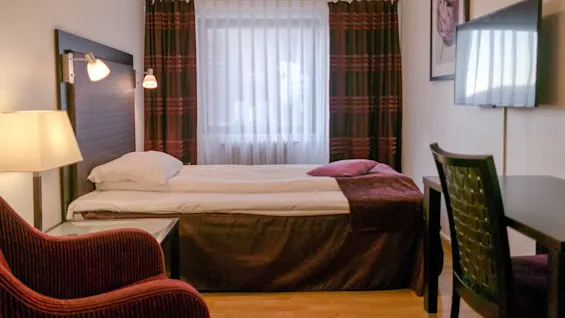 A small hotel room features a neatly made single bed with burgundy accents, a chair, desk, wall-mounted TV, and window with sheer curtains, creating a cozy ambiance.