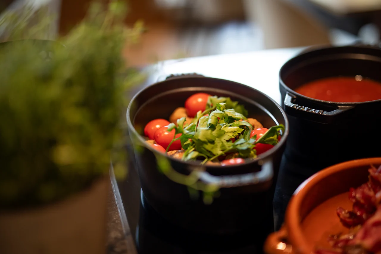 En svart gryte inneholder cherrytomater og bladsalat på en kjøkkenbenk. I nærheten står en annen gryte med rød saus, med en uskarp grønn plante i forgrunnen.