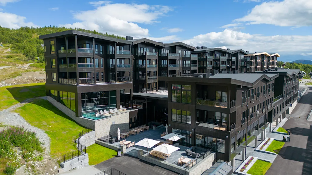 Et moderne, fler-etagers bygningskompleks med sorte facader står på en græsbeklædt skråning, med altaner og en rummelig terrasse. Omgivelser inkluderer klar himmel og nærliggende veje.